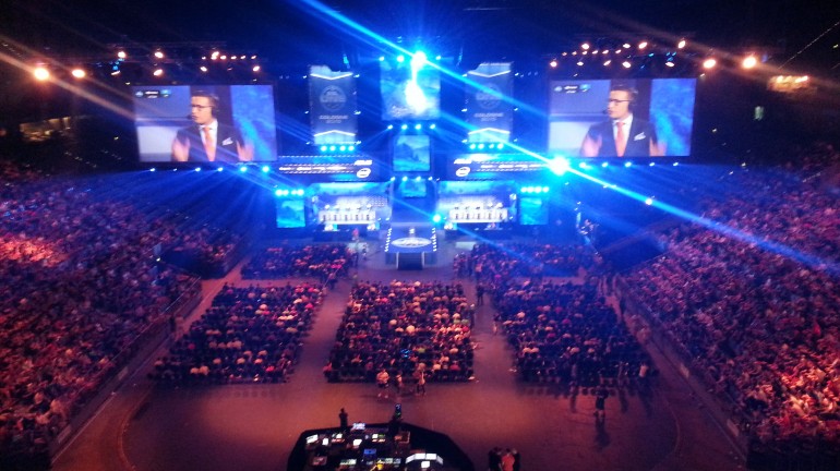 A bird's eye view of the LANXESS Arena. Production team at the back of the ground floor seating area. Casters on the screens doing pre-match discussions.